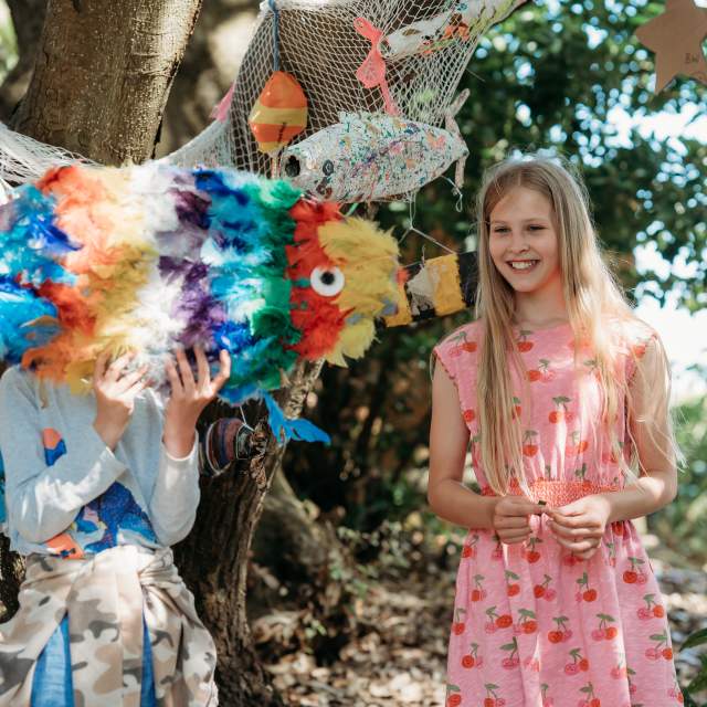 Two children are outdoors near trees. One child holds a colorful fish piñata, covering their face. The other child, smiling, wears a pink dress. The scene is playful and vibrant.