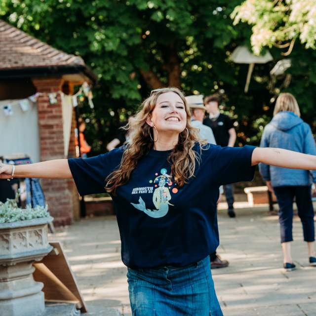 Woman joyfully extends arms in a sunny outdoor setting, wearing a dark shirt and jeans. She smiles with a bottle in hand. Others and greenery in background.