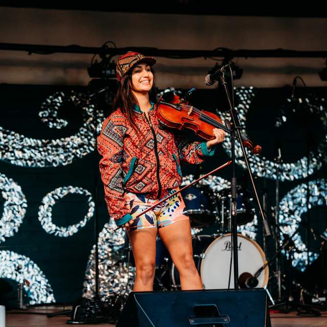 A performer on stage holding a violin, dressed in colourful, patterned attire, stands before an abstract backdrop of swirling designs, exuding joy and energy.