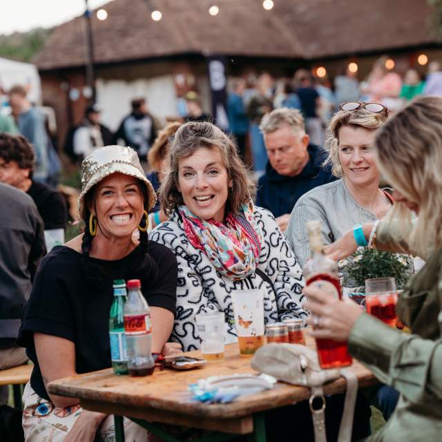 A lively outdoor gathering with smiling people at a wooden table. Drinks and snacks are visible, and a festive tent with lights is in the background.
