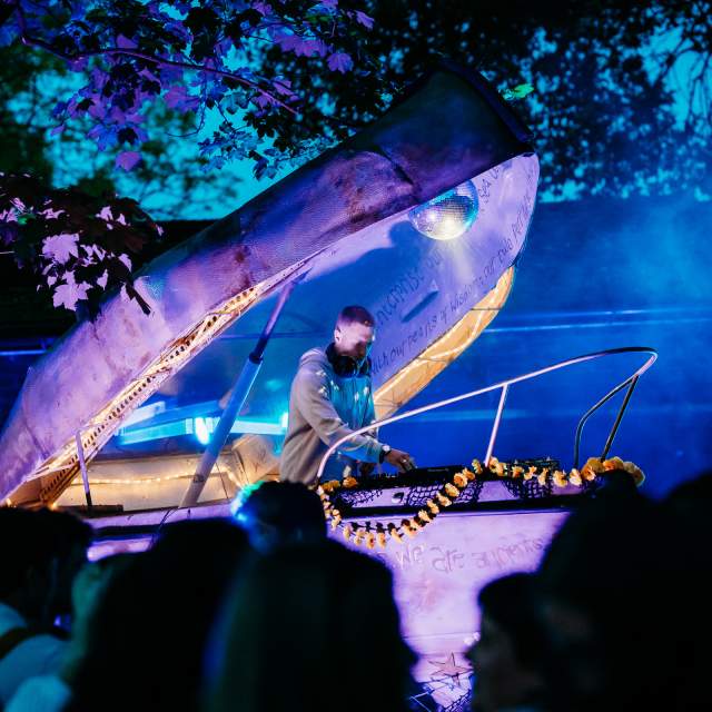DJ performing on a stage shaped like a giant open clam. The scene is bathed in vibrant blue and purple lights, creating a lively, festive atmosphere.