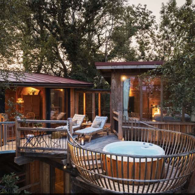 Treehouse retreat at sunset with large windows, cosy interior lighting, chaise lounges, and a hot tub on a wooden deck surrounded by lush trees.