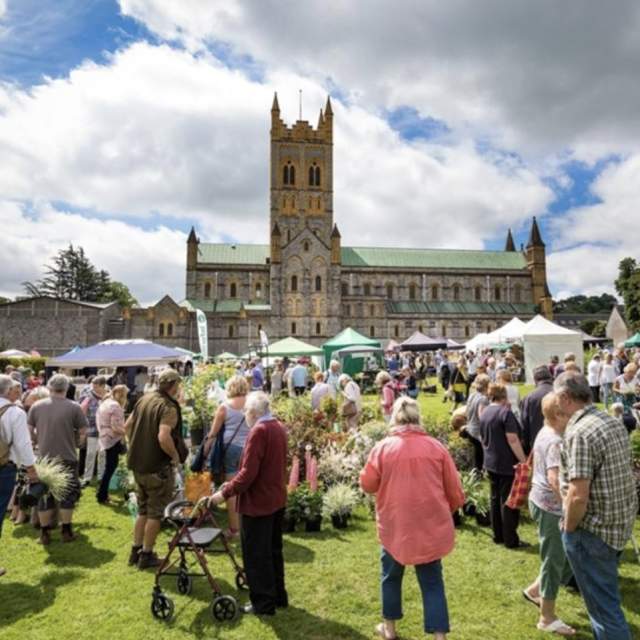 Crowded outdoor garden festival in front of a large, historic cathedral. People browse plant stalls under partly cloudy skies, conveying a lively atmosphere.