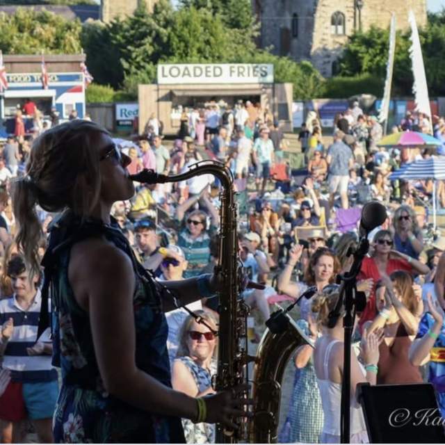 A female saxophonist performs on stage, silhouetted against a lively crowd at an outdoor festival. Attendees clap and cheer under a sunny sky, exuding joy.