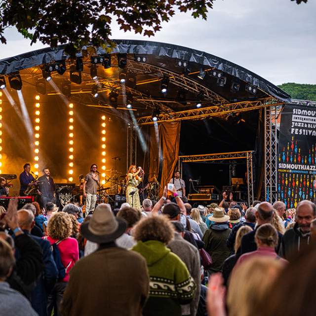 Outdoor music festival scene with a lively crowd watching performers on a lit stage. Banners read "Sidmouth Jazz and Blues Festival." Trees frame the view.