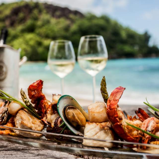 A plate of assorted seafood, including shrimp and mussels, on a table with two glasses of white wine. A scenic beach and lush greenery in the background.