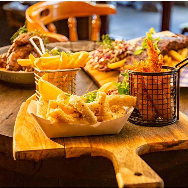 A rustic table holds a wooden board with crispy fries in a metal basket, calamari, and ribs with garnish. The scene is warm, cosy, and inviting.