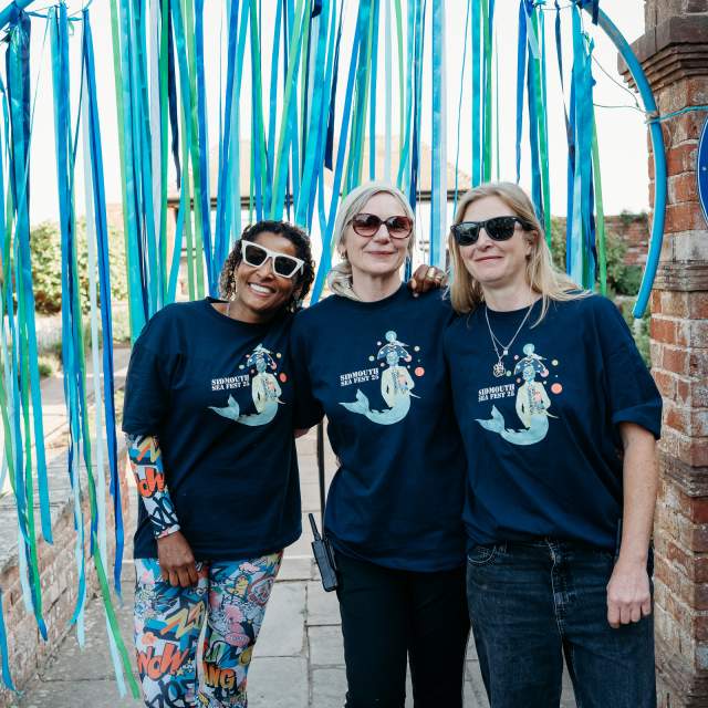 Three people wearing matching blue shirts and sunglasses stand smiling under blue and green streamers in an outdoor setting, conveying a joyful mood.