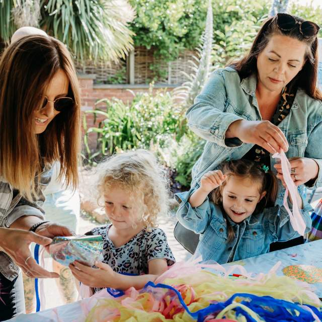Two women and two children engage in a colourful craft activity outside, surrounded by bright materials. The scene is lively and cheerful, set against a garden backdrop.