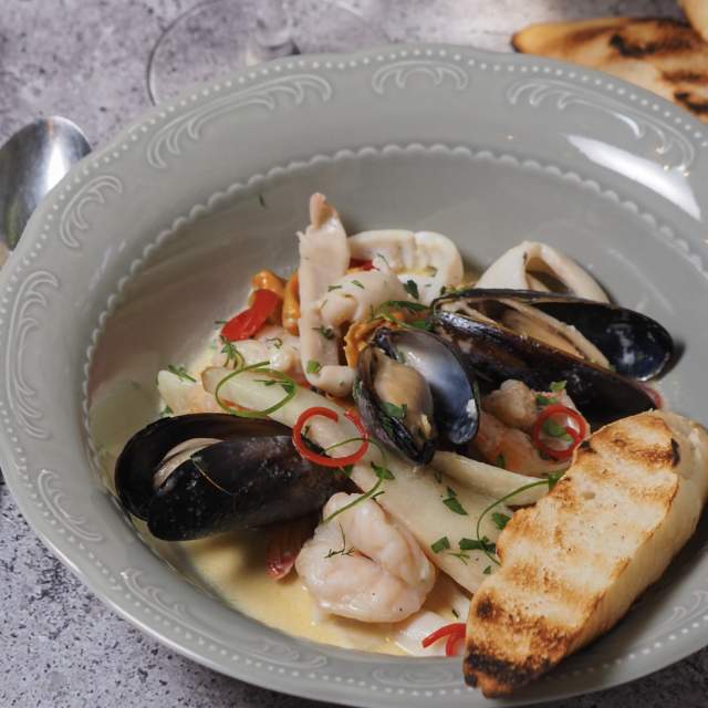 A grey plate featuring a seafood stew with shrimp, mussels, and calamari, garnished with herbs and chilli slices. Grilled bread is on the side. Casual dining setting.