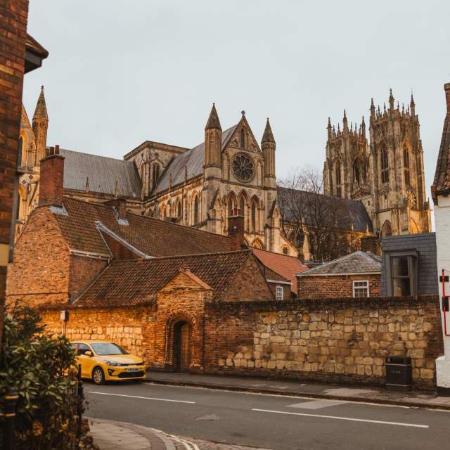 Beverley Minster towering in the background with its Gothic spires, viewed from a historic cobbled street lined with old brick houses in the foreground