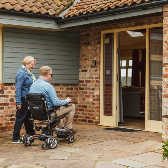 2 people 1 in motorized wheelchair approaching a cottage door