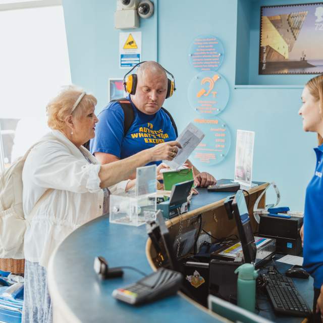 2visitors at Reception Desk, with 1 person behind the counter. One person is holding a leaflet, one is wearing noise cancelling headphones.