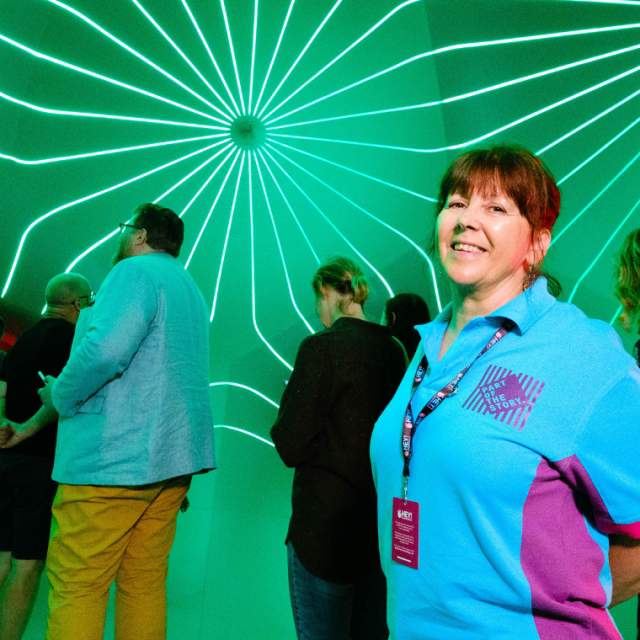 A hey volunteer at an event, with people in the background along with a bright green neon wall.
