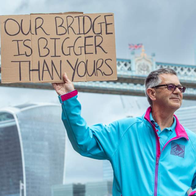 A HEY Volunteer in his iconic blue uniform stood in front of Tower Bridge in London holding a sign which says our bridge is better than yours, referring to the Humber Bridge in East Yorkshire.