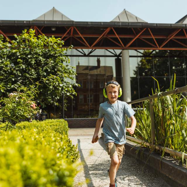 Little boy running through Hull Museums Quarter gardens  towards the camera wearing noise cancelling headphones