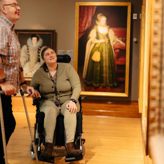 2 people view painting in art gallery 1 person on crutches 1 person in motorized wheelchair