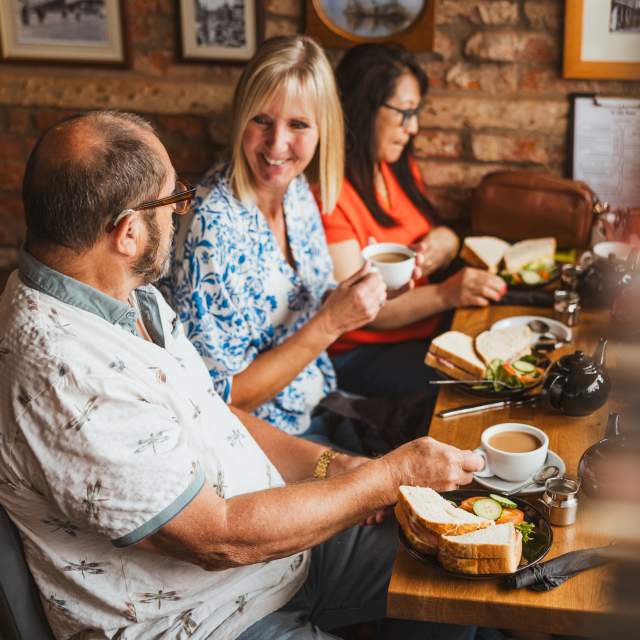 3 People sat a a table enjoying coffee and sanwichiches.