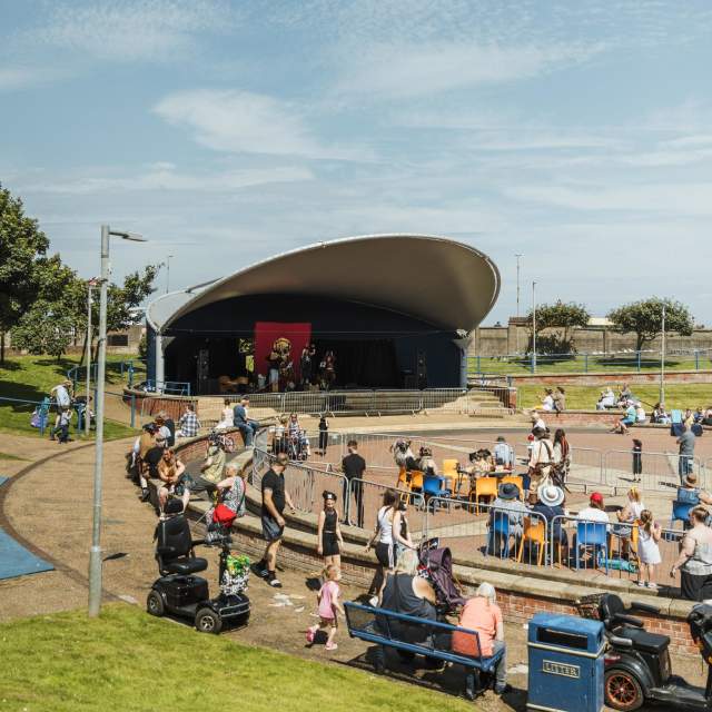 Wide view of stage and viewing area with crowd of people, benches and motorised wheelchairs