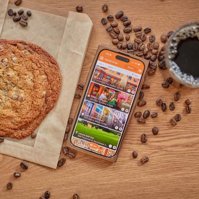 An overhead view of a table topped with a cookie, a cup of coffee, coffee beans scattered, and a cell phone with the Galena Country app open.