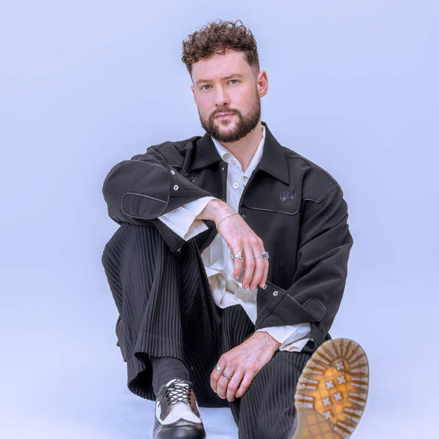 Calum Scott sits in front of a lilac backdrop. He is wearing a black denim overshirt over a loose fitting white shirt plus pinstriped black trousers and Doc Marten loafers. He is sat with his arm resting on his right knee and his left foot out straight so you can see to sole of his shoe.