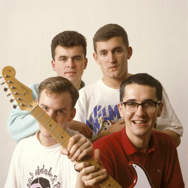 The Housemartins are pictured in front of a beige backdrop. Paul Heaton and Norman Cook are sat in the forgroung. Heaton is wearing a red shirt and holding a guitar which is covering Cook's face. Cook is wearing a white t-shirt with a black graphic printed on it. behind them Dave Hemingway and Stan Cullimore are stood above and are wearing a blue sweatshirt and white over sized t-shirt respectively.