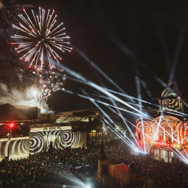 An overview of Hull City Hall and the Maritime Museum, lit up at night with spirals and flashlights, whilst fireworks explode overhead. A large crowd is gathered to watch the display.