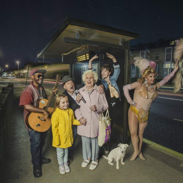An older lady with a big beaming smile stands at a bus stopped surrounded by performers, a dancer, guitar player, dog and a little girl in a yellow coat.