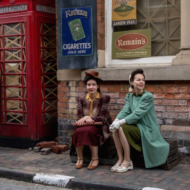 Actors dressed in 1940s clothing sit beside a vintage British red telephone box on a cobbled street, surrounded by period military uniforms and retro advertising signs.