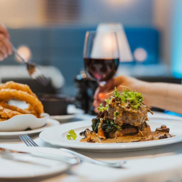A steak dinner is laid out on a crisp whie table cloth. The fillet steak in the foreground is garnished with mixed greens. Large crispy onion rings are in a bowl to the left and a fork is reaching to pick one up. Another hand is picking up a large glass of red wine. The restaurant in the background is out of focus but the seating is dark blue and candles are flickering in the background.