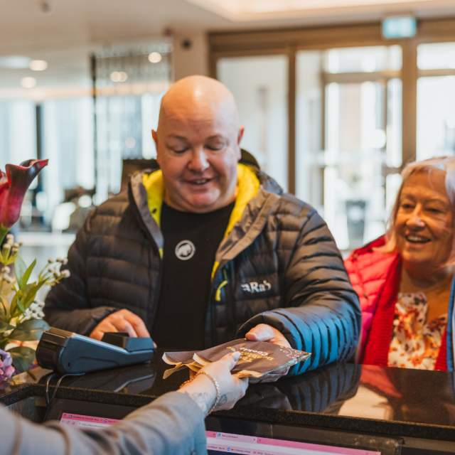 Guests checking in at a hotel reception desk, with staff assisting and travel brochures, flowers and luggage visible in a bright, modern hotel lobby.