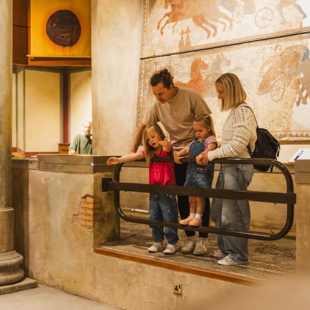 A family with two young children stands in the Hull & East Riding Museum, looking over a railing at an exhibit, with a large ancient mural depicting horses and chariots on the wall behind them.