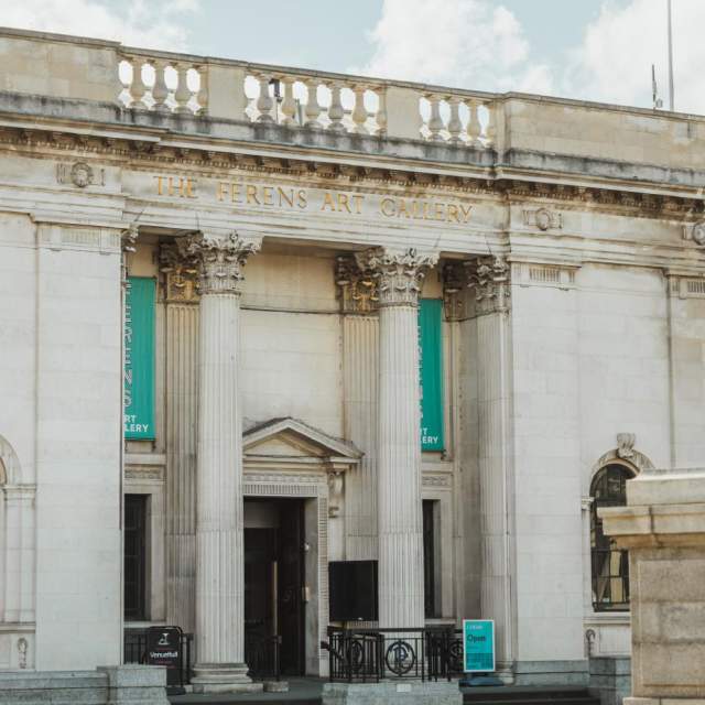 Front exterior of the Ferens Art Gallery in Hull, showing its neoclassical stone façade with tall columns, decorative carvings and entrance banners on a bright day, with the nearby historic lamp feature in the foreground.