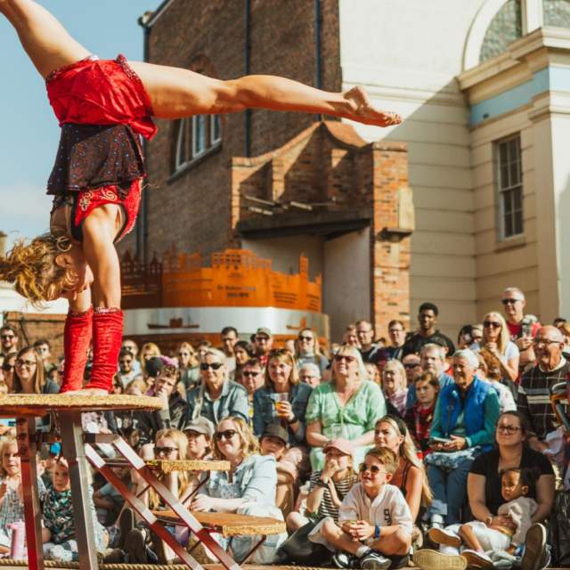 A woman in a red leotard is performing a hand stand on a raised platform with a crowd surrounding her.