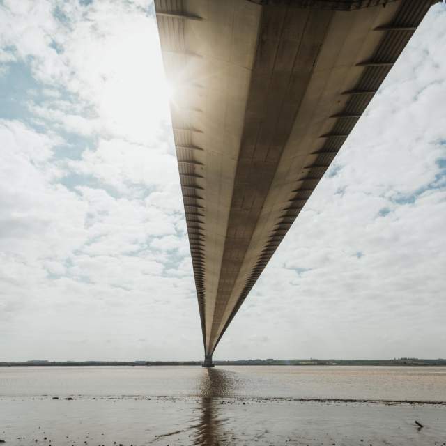 A view beneath the Humber bridge, with clouds either side and the foreshore just in front of the viewer.