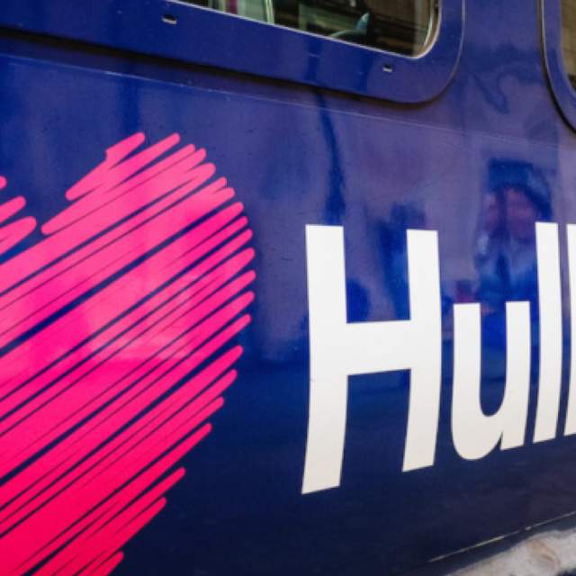 Close-up of a Hull Trains carriage displaying the company logo, featuring a large pink heart graphic and white “Hull Trains” branding on a dark blue train exterior at a station platform. Reflections of passengers and station buildings are visible in the train windows.