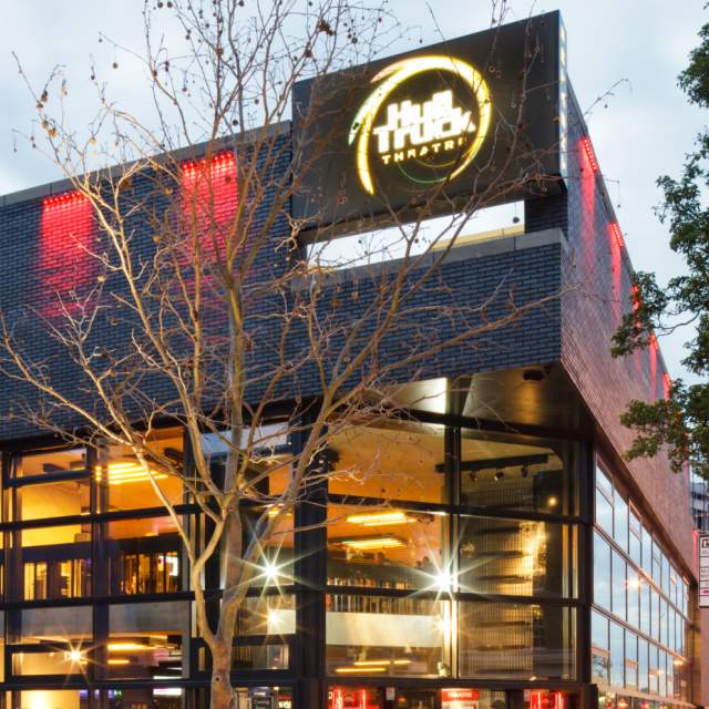 Exterior view of Hull Truck Theatre at dusk, showing the modern brick building illuminated with red lighting and large glass windows, with the theatre’s bright circular logo displayed above the entrance and trees framing the scene.