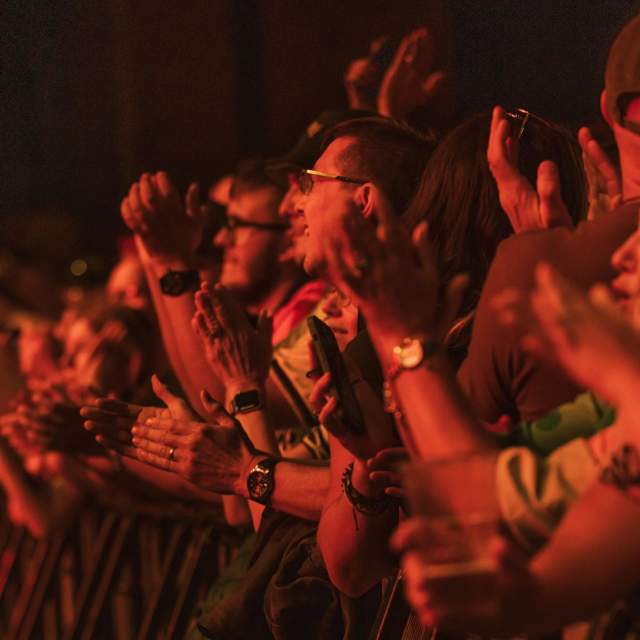 Conceert goers at Hull's Zebedee's Yard applauding and looking up at the stage. The lights from the stage are reflecting back at them washing the crowd with a red hue.