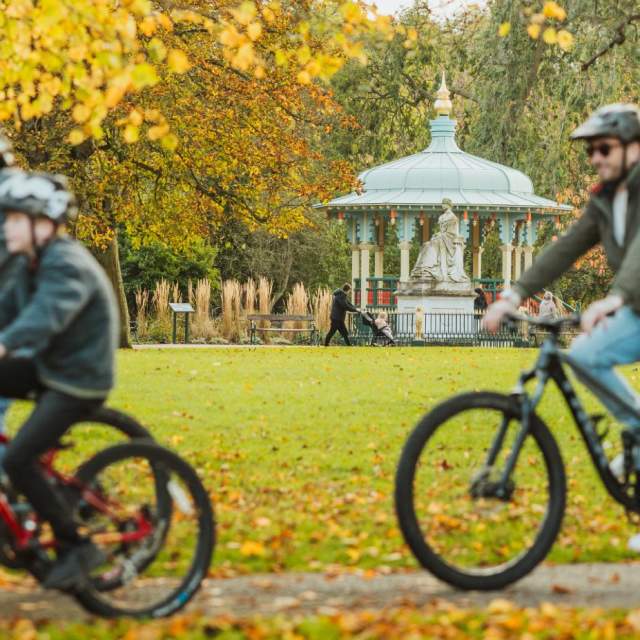 Cyclists riding along a tree‑lined path in Hull’s Pearson Park during autumn, with the bandstand visible in the background