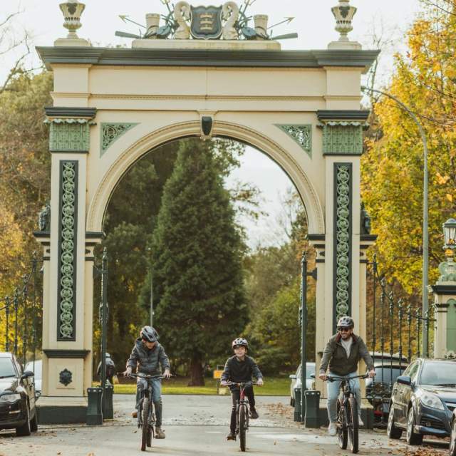Cyclists riding through the ornate entrance archway of Hull’s Pearson Park, surrounded by parked cars and autumn trees
