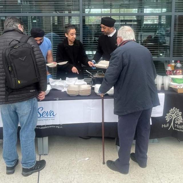 Sikhs in Hull stand behind a buffet table giving out food to gentleman at Hull History Centre.