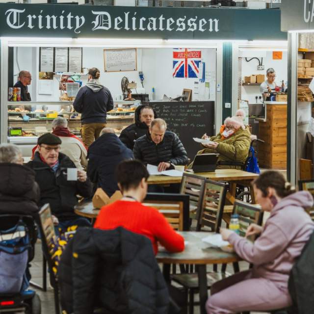 Visitors, including wheelchair users and people with mobility aids, enjoy the accessible indoor food stalls and seating areas at Trinity Market in Hull. The scene highlights step‑free access, spacious walkways, and inclusive dining options within the market