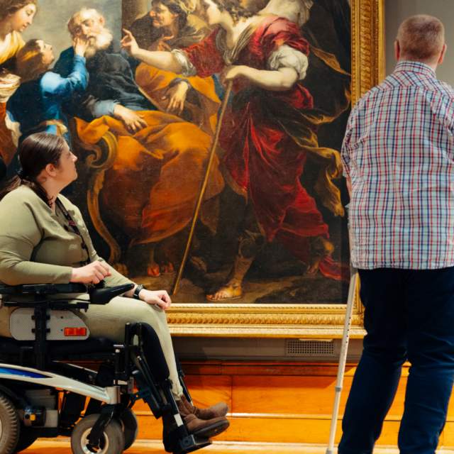 A powered wheelchair user and another visitor with a mobility aid admire the  large classical painting 'Tobias Healing the Blind Tobit' (c.1650) by Valerio Castello in the spacious, accessible Ferens art gallery Hull .