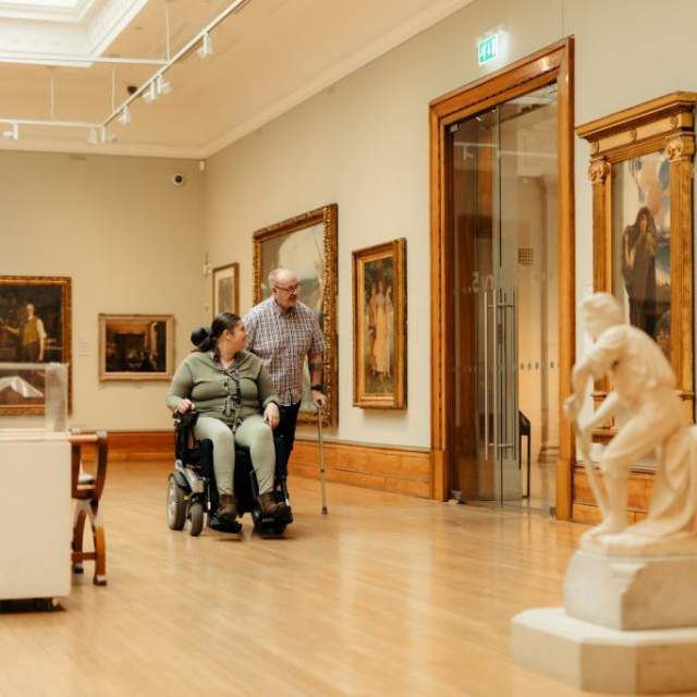 A wheelchair user explores Ferens art gallery Hull with another visitor, surrounded by sculptures and framed artwork. The scene highlights inclusive museum accessibility