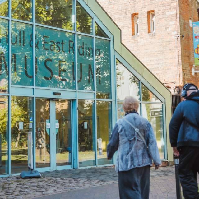 Two visitors approach the entrance to Hull & East Riding Museum, a brick-built museum with large glass doors, clear signage and a level pathway leading to the accessible entrance."