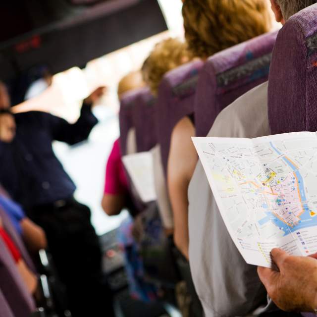 Group of passengers seated on a coach while following a printed map, with a guide speaking at the front during a sightseeing tour.