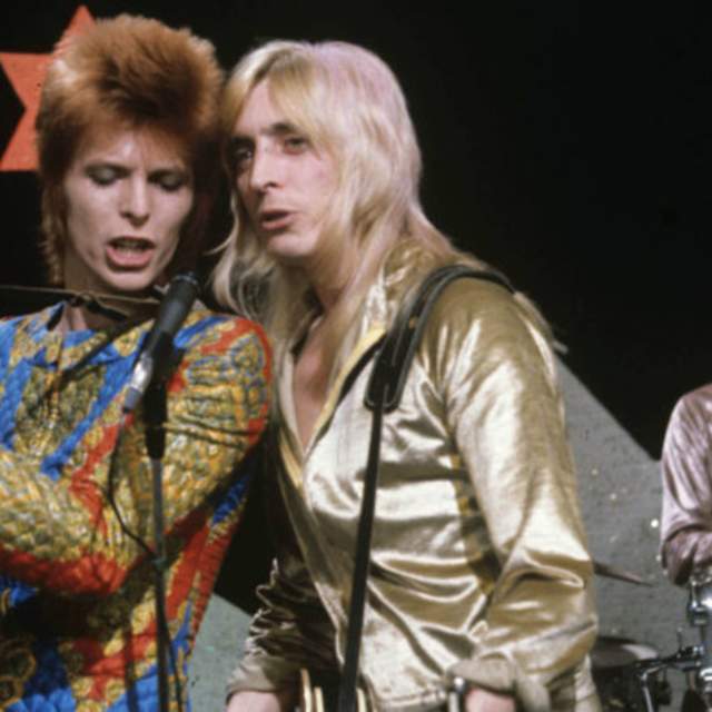 David Bowie and Mick Ronson singing into microphones on stage. They are in classic 1970s outfits complete with volumious hair. Bowie is in a yellow, red and blue jump suit with a ginger mullet, he is playing a black guitar. Ronson has platinum long blonde hair and wears a gold shiny shirt. Woody Woodmansey plays shiny silver drums in the background, he wears a glittery pink top and has long brown curly hair.