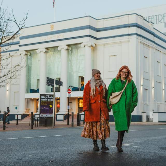 Hull New Theatre in the winter, the white and blue building is light by vertical lights up the colums. Two ladies are walking away after seeing a show. They are bundled up in hats and coats. The lady on the left has blonde hair and is wearing a rusy red fur coat and a beige hat. The lady on the right is wearing a green coat and has red hai and glasses.