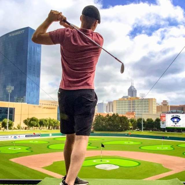 A man playing golf at Victory Field baseball field, with the Indy skyline in the background