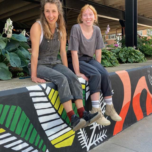 Two female artists sit atop a mural painted in Houghton